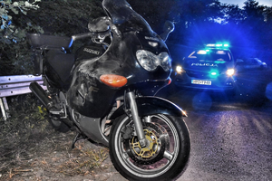 stojący przy metalowej barierce motocykl marki Suzuki, w tle policyjny radiowóz z włączonymi sygnałami świetlnymi